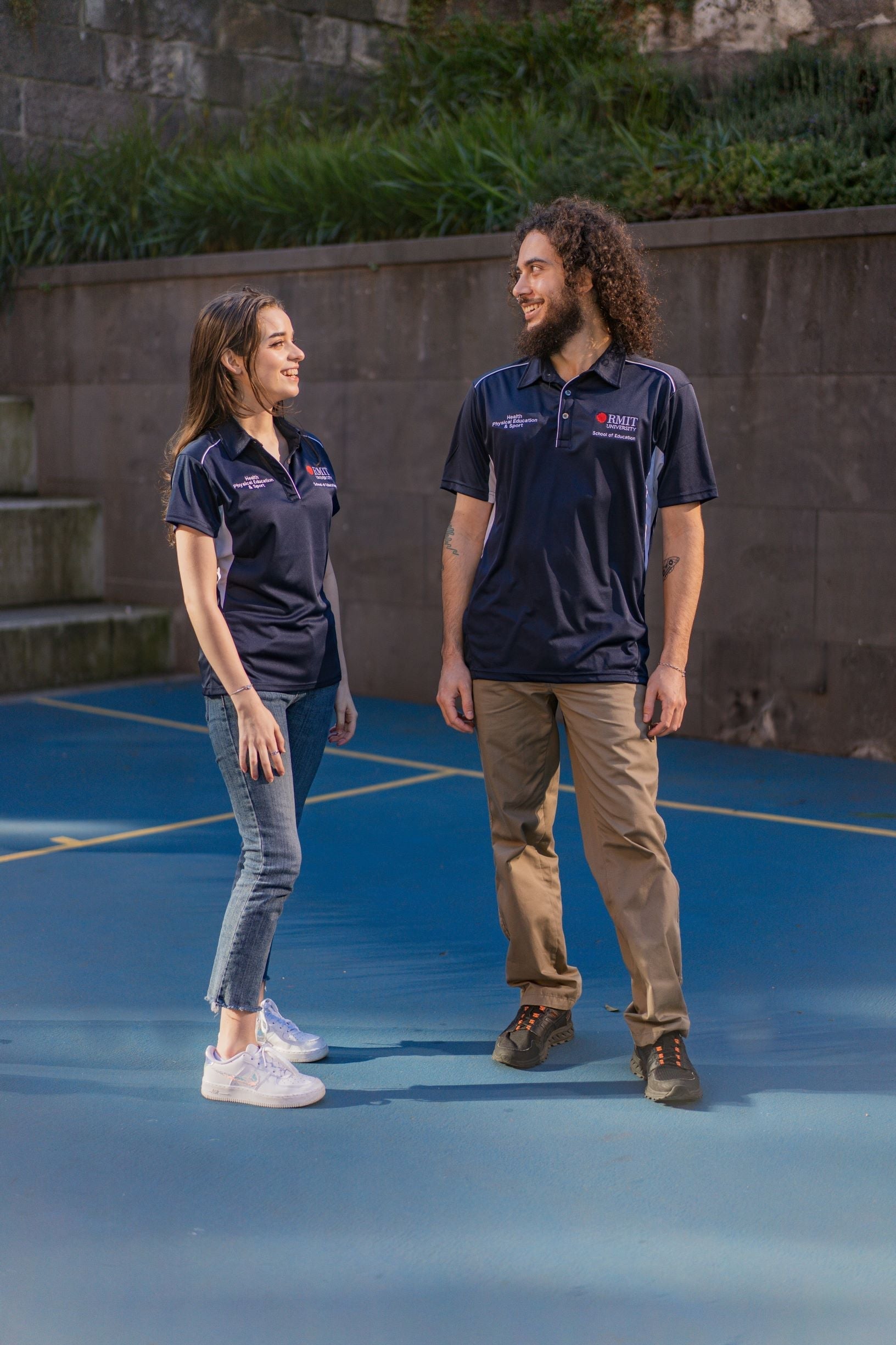 Male and female model on campus looking at each other while wearing the RMIT Store Health, Physical Education & Sport Uniform Polo. The navy and white polyester polo features the RMIT University logo in red and white on the left chest, with “Health, Physical Education & Sport” in white on the right chest. Both models are outdoors on campus.